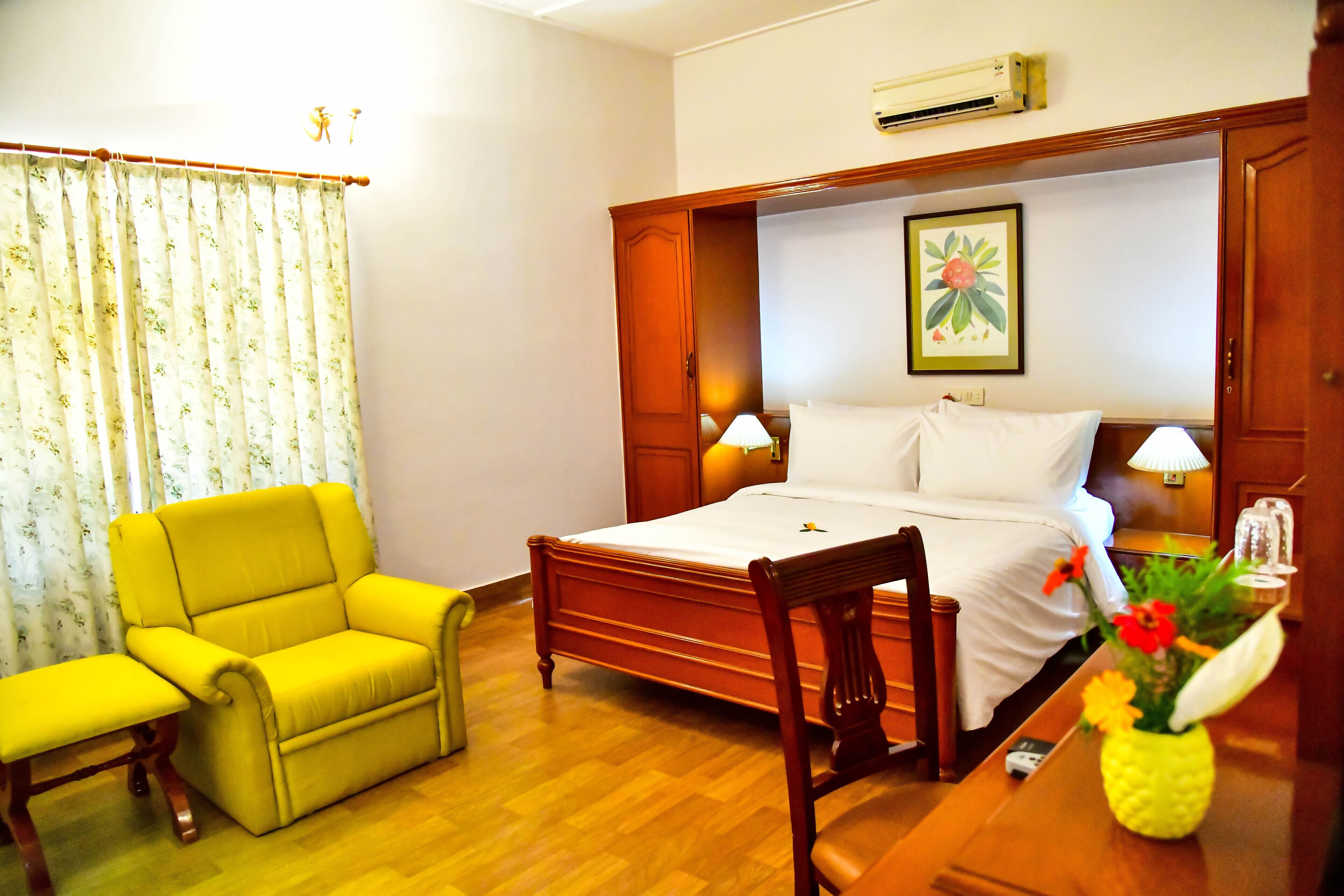 A hotel room with a bed, a yellow armchair, a wooden nightstand, and a wall-mounted air conditioner.