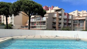 A swimming pool with a tiled deck, surrounded by a white fence. In the background, there are multi-story buildings with balconies and a tree. - Renovated Studio in Cap d'Agde by the Beach (Cap d'Agde)