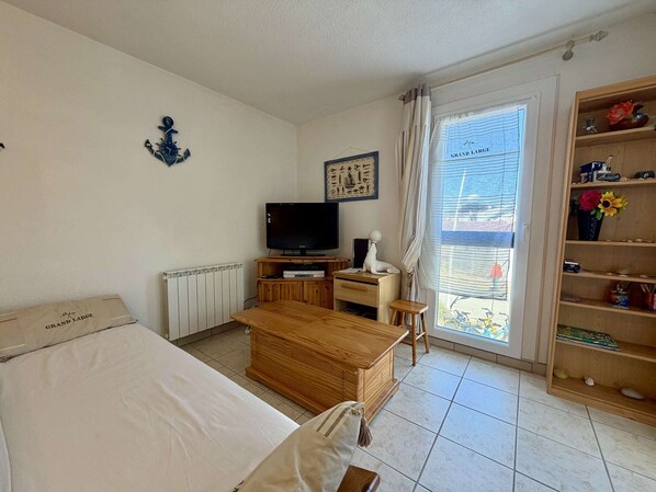 Habitación de hotel con cama, escritorio de madera, televisor, radiador, ventana con cortinas y estantería.