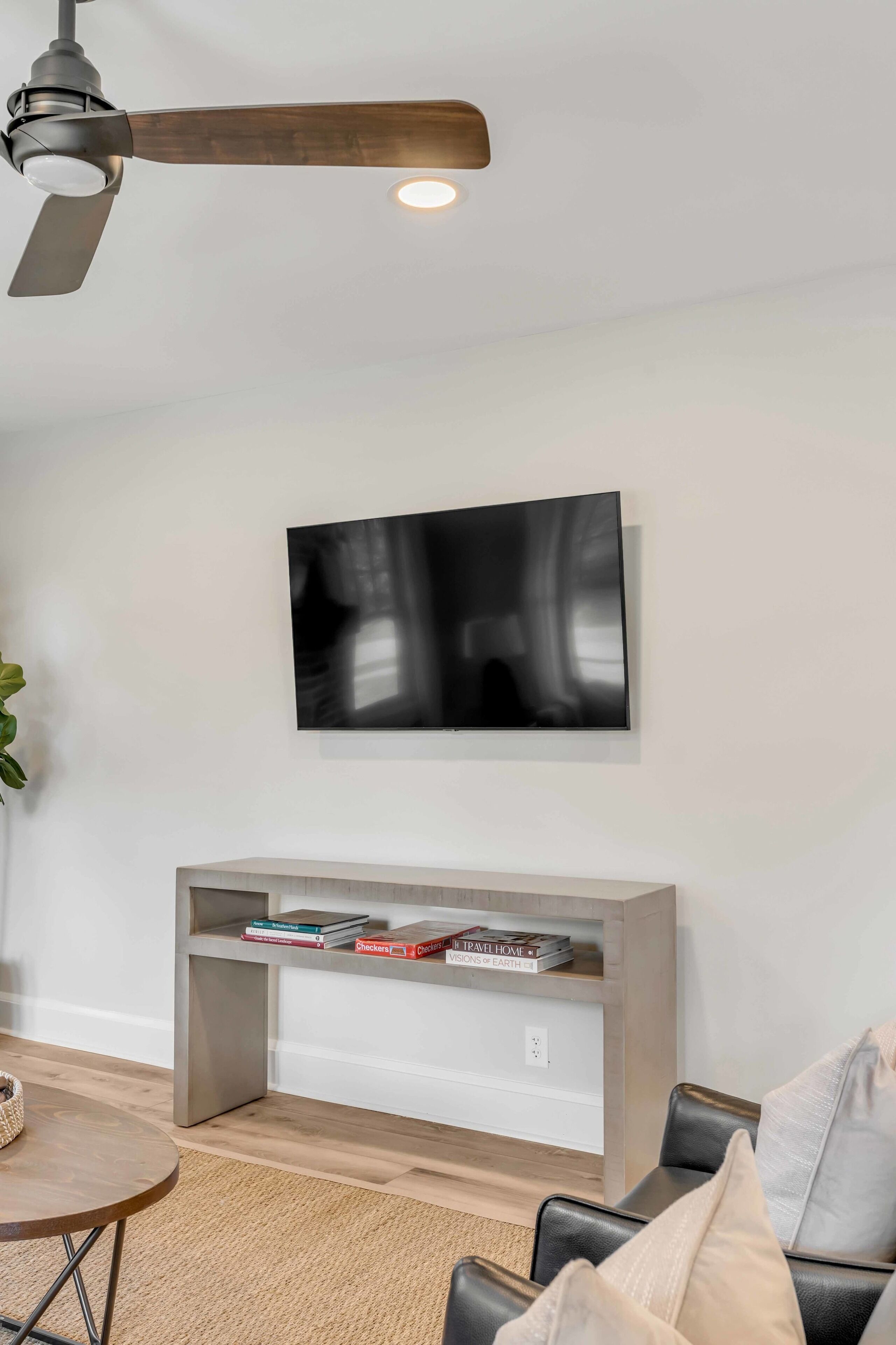 A modern living room with a ceiling fan, a mounted flat-screen TV, a built-in bookshelf, a round coffee table, and a black leather sofa.