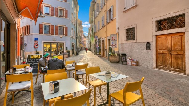 Una calle adoquinada con mesas al aire libre, edificios con contraventanas y un cielo despejado.