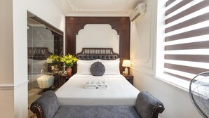 Un dormitorio ordenado con una cama grande, una mesita de noche con lámpara, un pequeño rincón de descanso y vistas al exterior a través de una ventana con persianas.