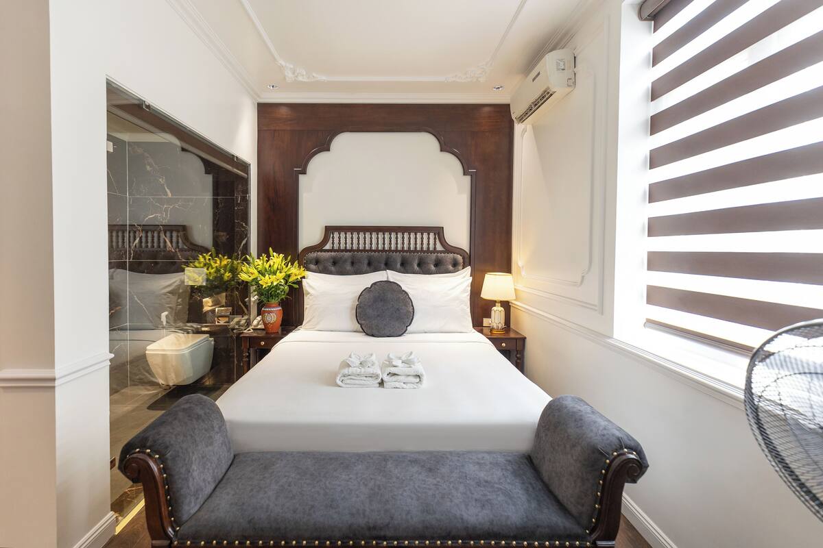 A neatly arranged bedroom with a large bed, a bedside table with a lamp, a small seating area, and a view of the outside through a window with blinds.