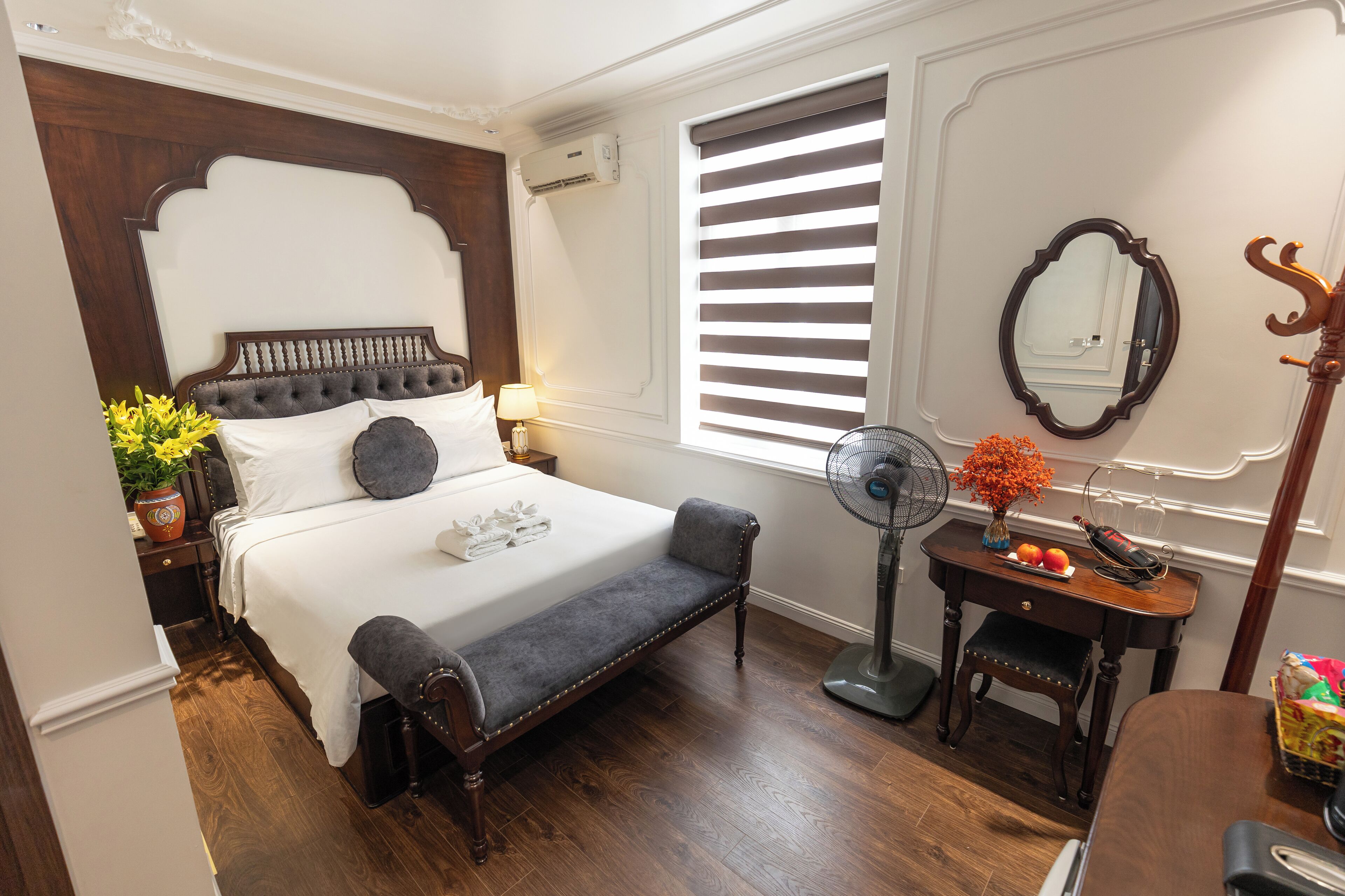 A bedroom with a large bed, bedside table, a bench, a dresser, a mirror, and a window with blinds.