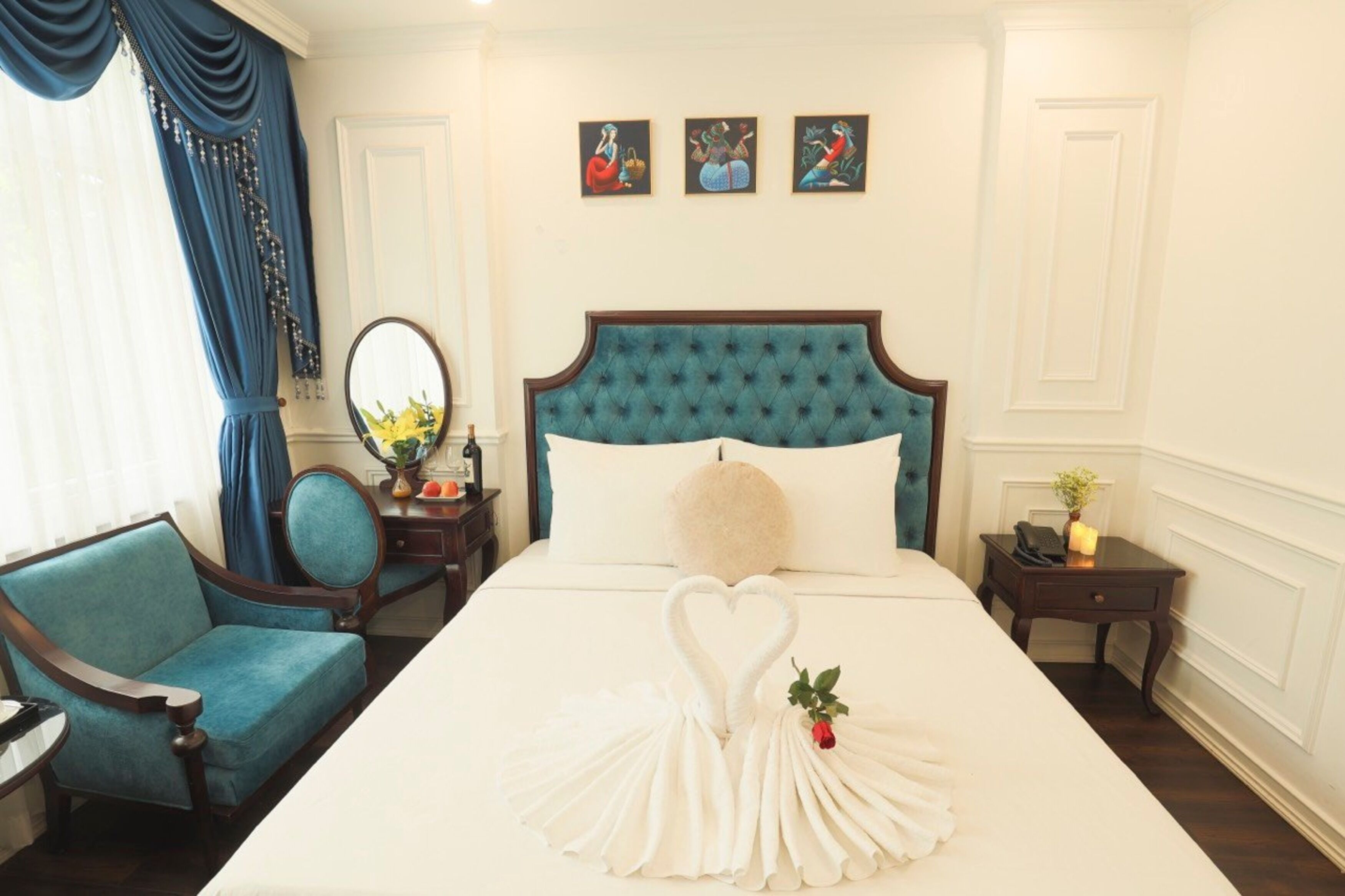 A hotel room with a large bed, a blue upholstered chair, a bedside table with a lamp, and a wall with three framed artworks.