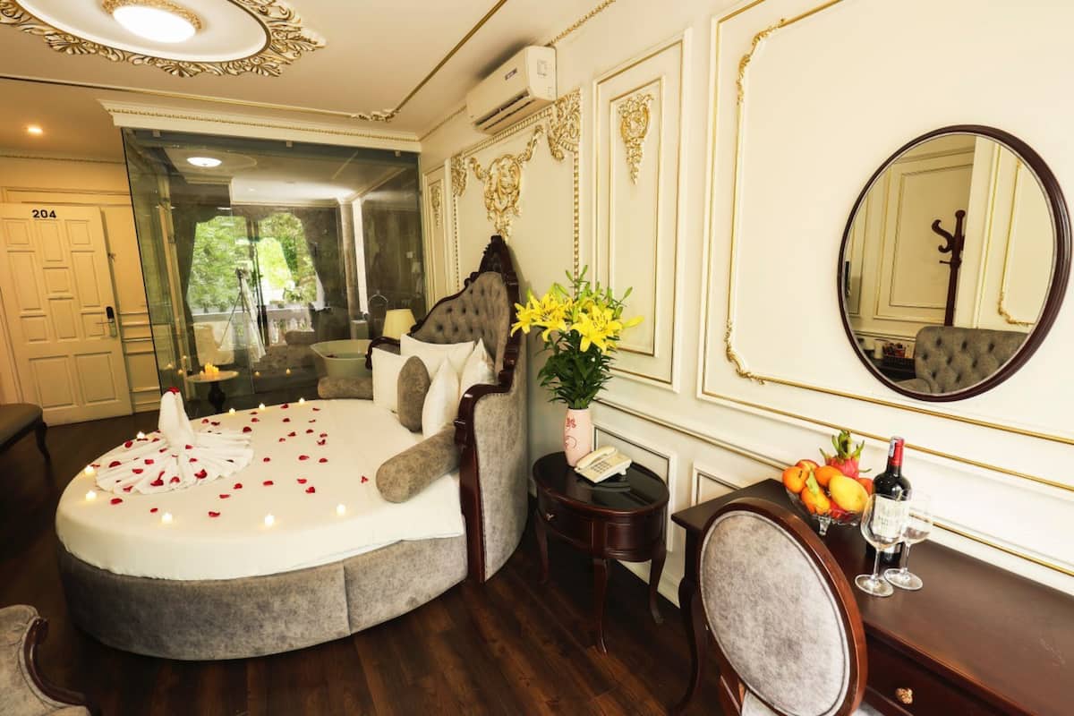 A luxurious bedroom with a round bed, a mirror, a vase with flowers, a fruit bowl, and a bottle of wine.