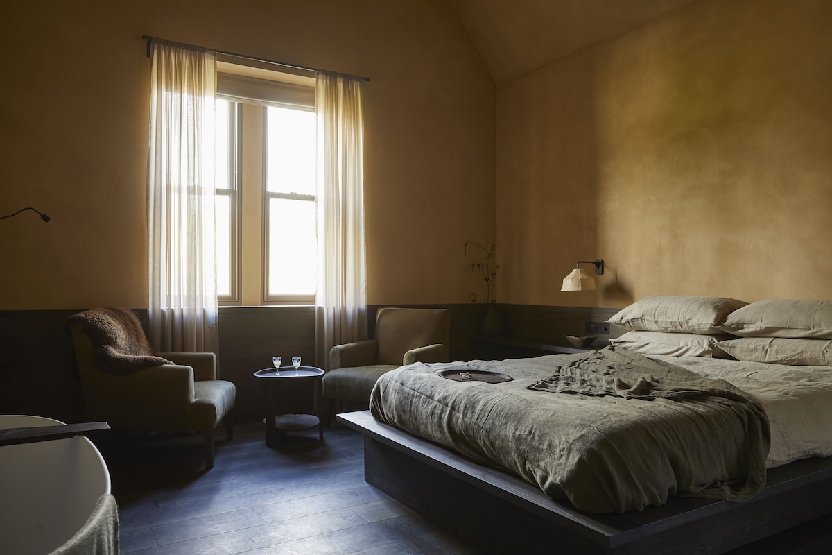 Een slaapkamer met een groot bed, een stoel, een kleine tafel en een raam met gordijnen.