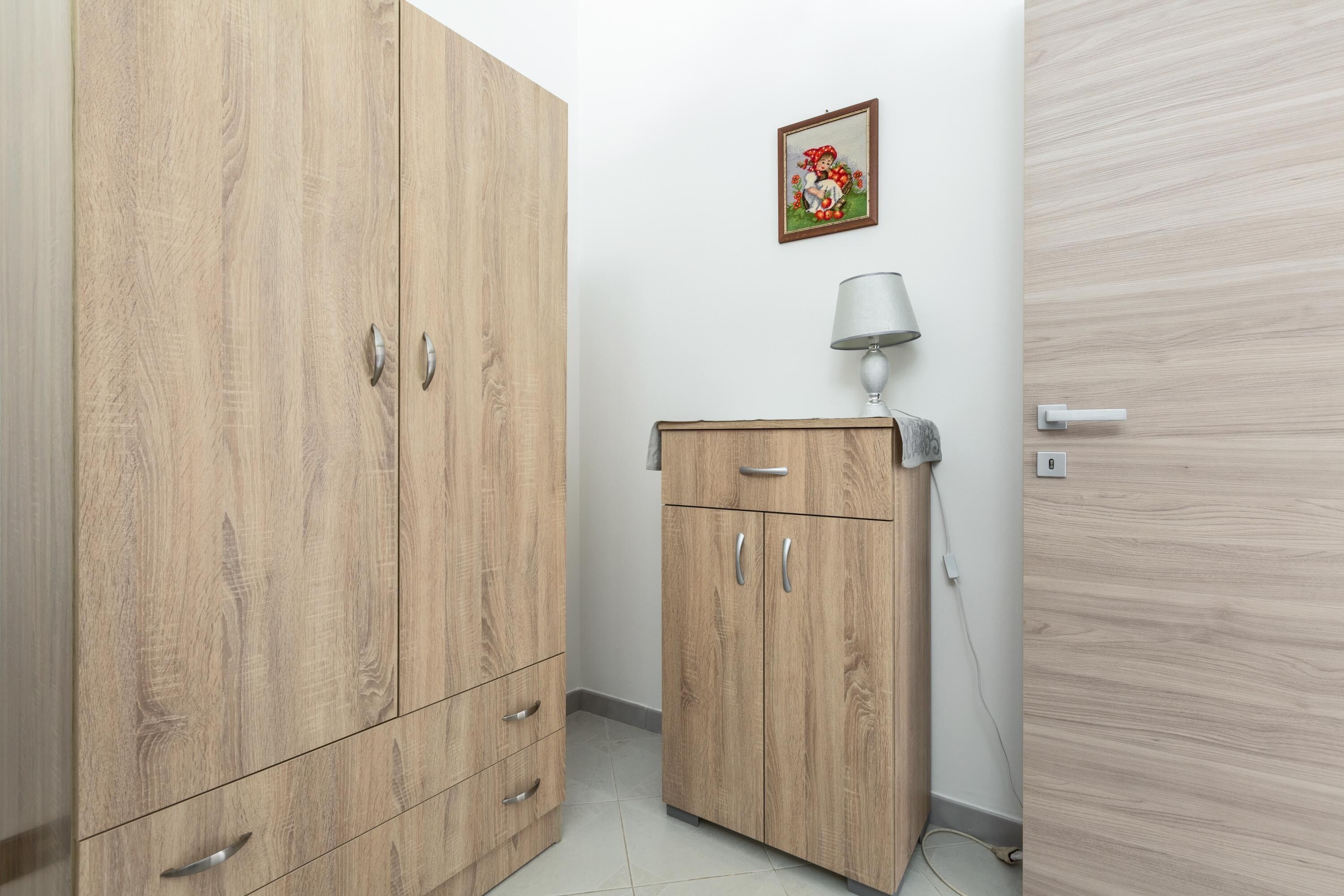 A room with a wooden wardrobe, a small cabinet, and a wall-mounted lamp.