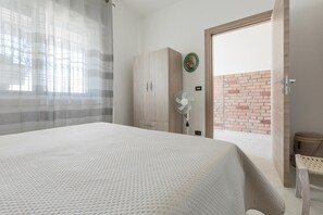 A bedroom with a bed, a wardrobe, a fan, and a view of a brick wall through an open door. - 3825 Sabbiabianca - Aptm B by Barbarhouse (Porto Cesareo)