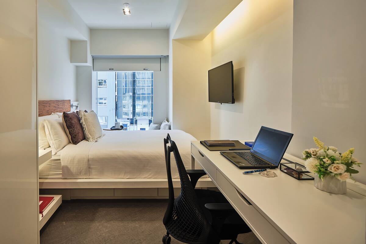 A modern hotel room with a bed, a desk with a laptop, a chair, a TV mounted on the wall, and a window with a city view.