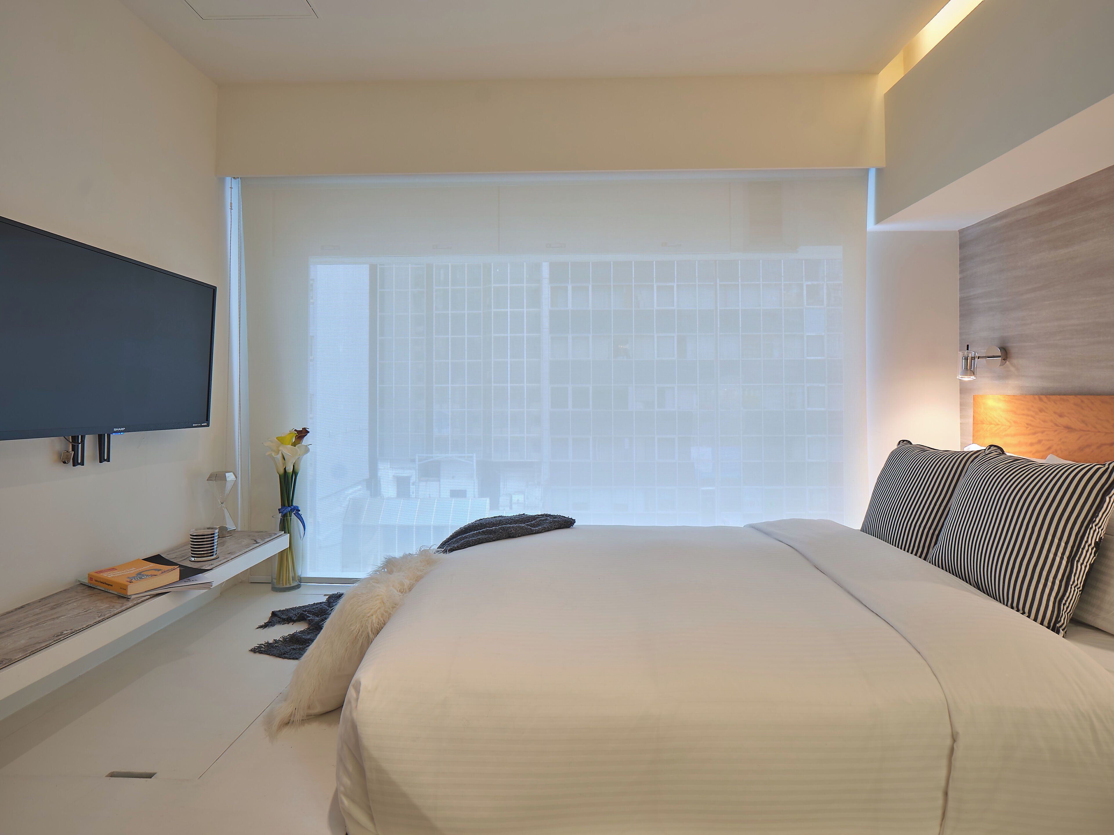 A modern hotel room with a large bed, a flat-screen TV mounted on the wall, a bedside table with a lamp, and a view of the city through large windows.