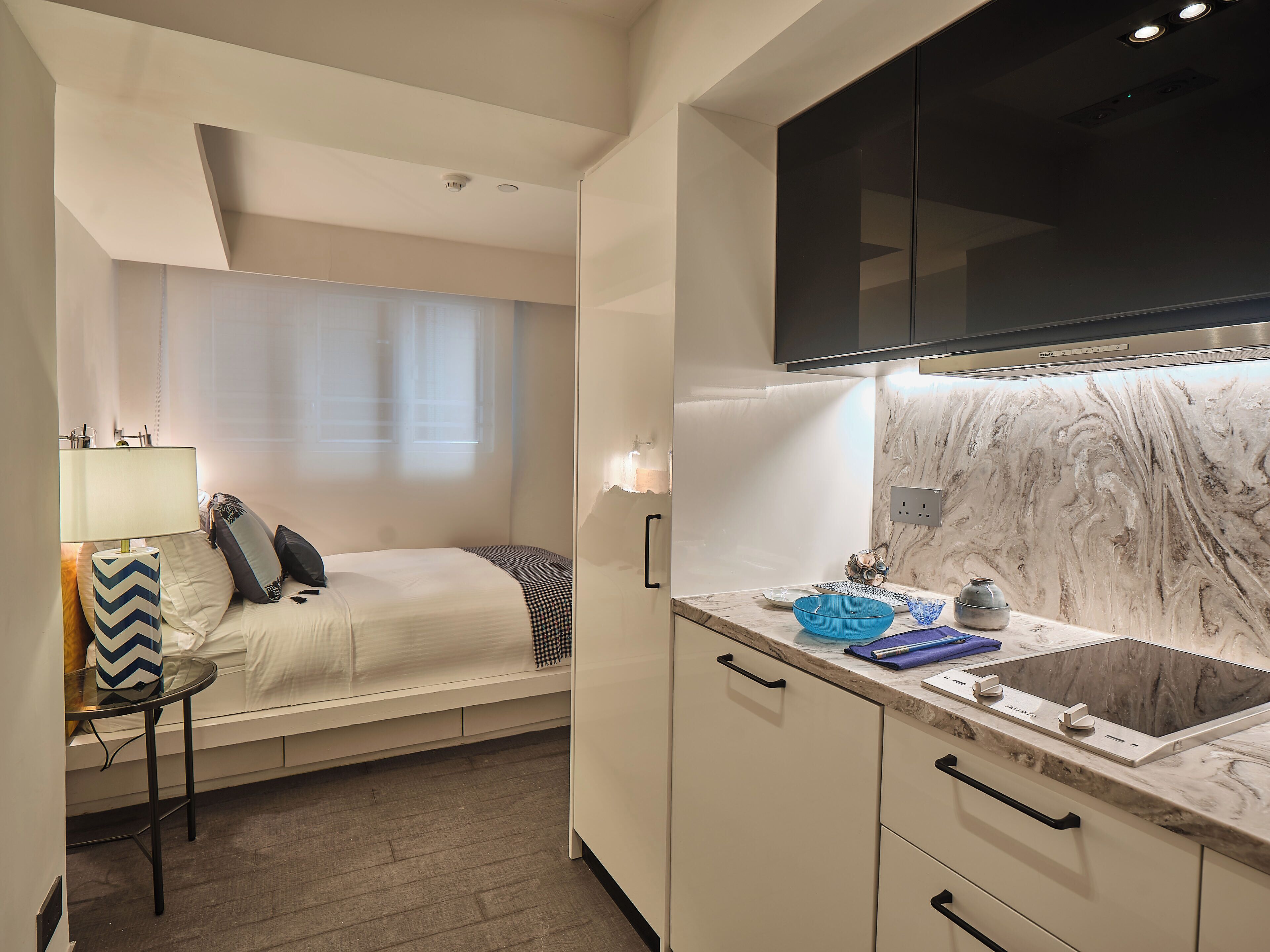 A compact hotel room with a bed, bedside table, and a small kitchenette with a sink and countertop.