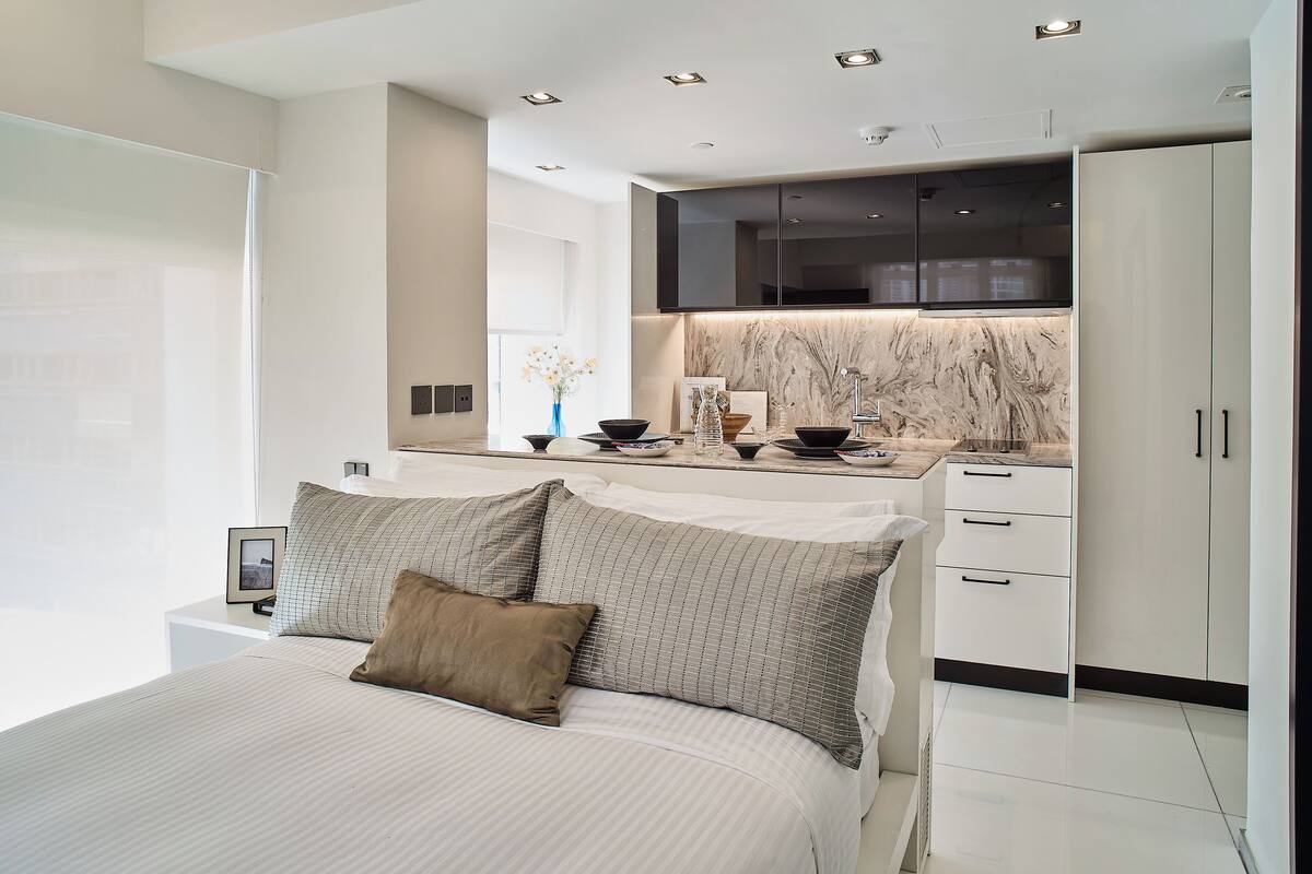 A modern bedroom with a large bed, a bedside table, and a view of a kitchen area.