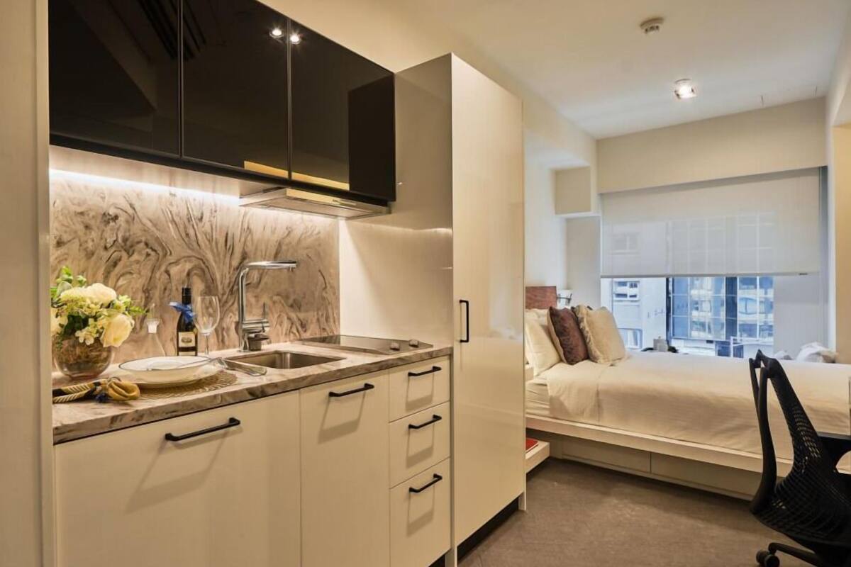 A modern hotel room with a kitchen area, a bed with a city view, and a black office chair.