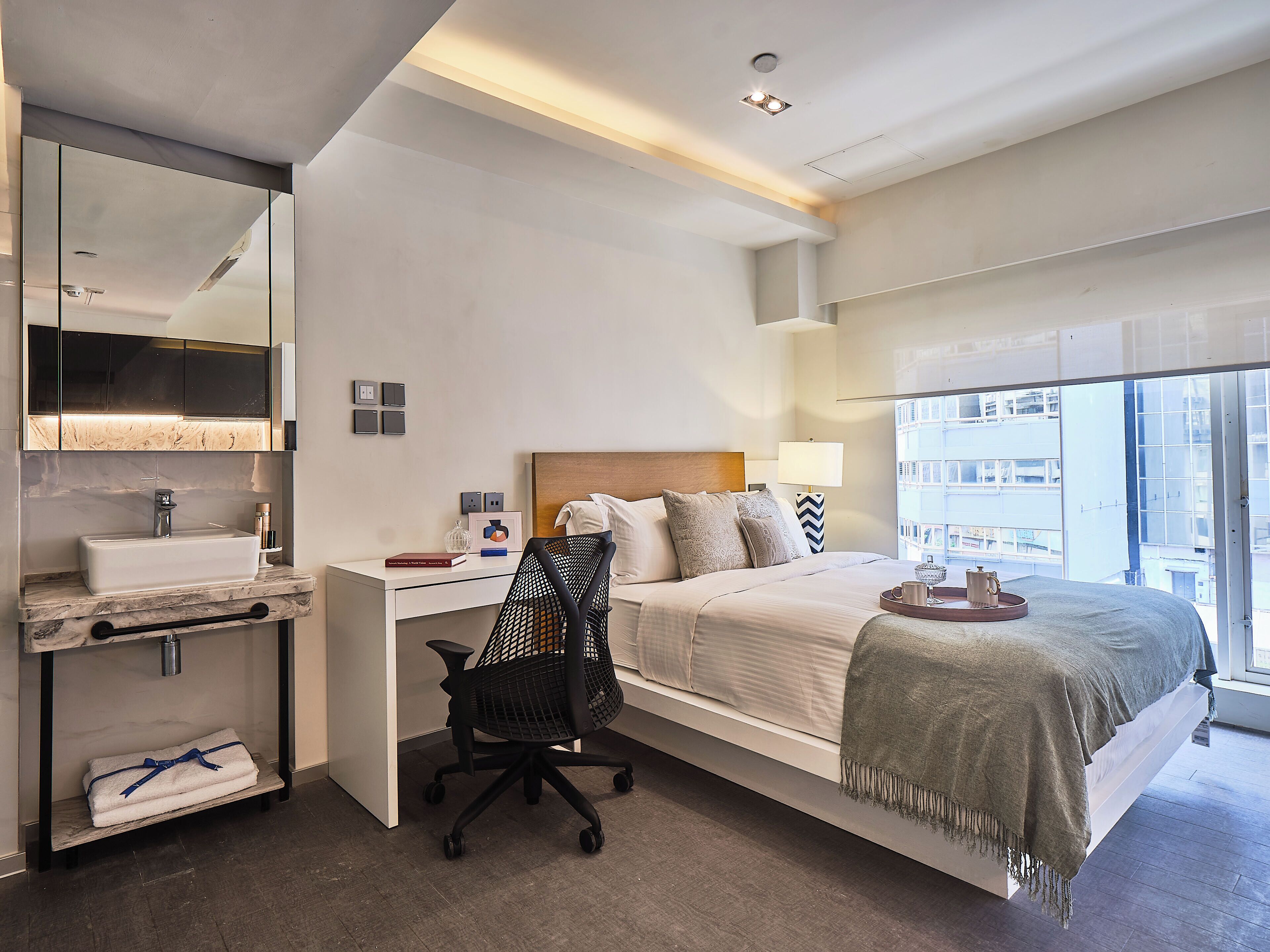 A modern hotel room with a large bed, a desk with a sink, a chair, and a view of the city through large windows.