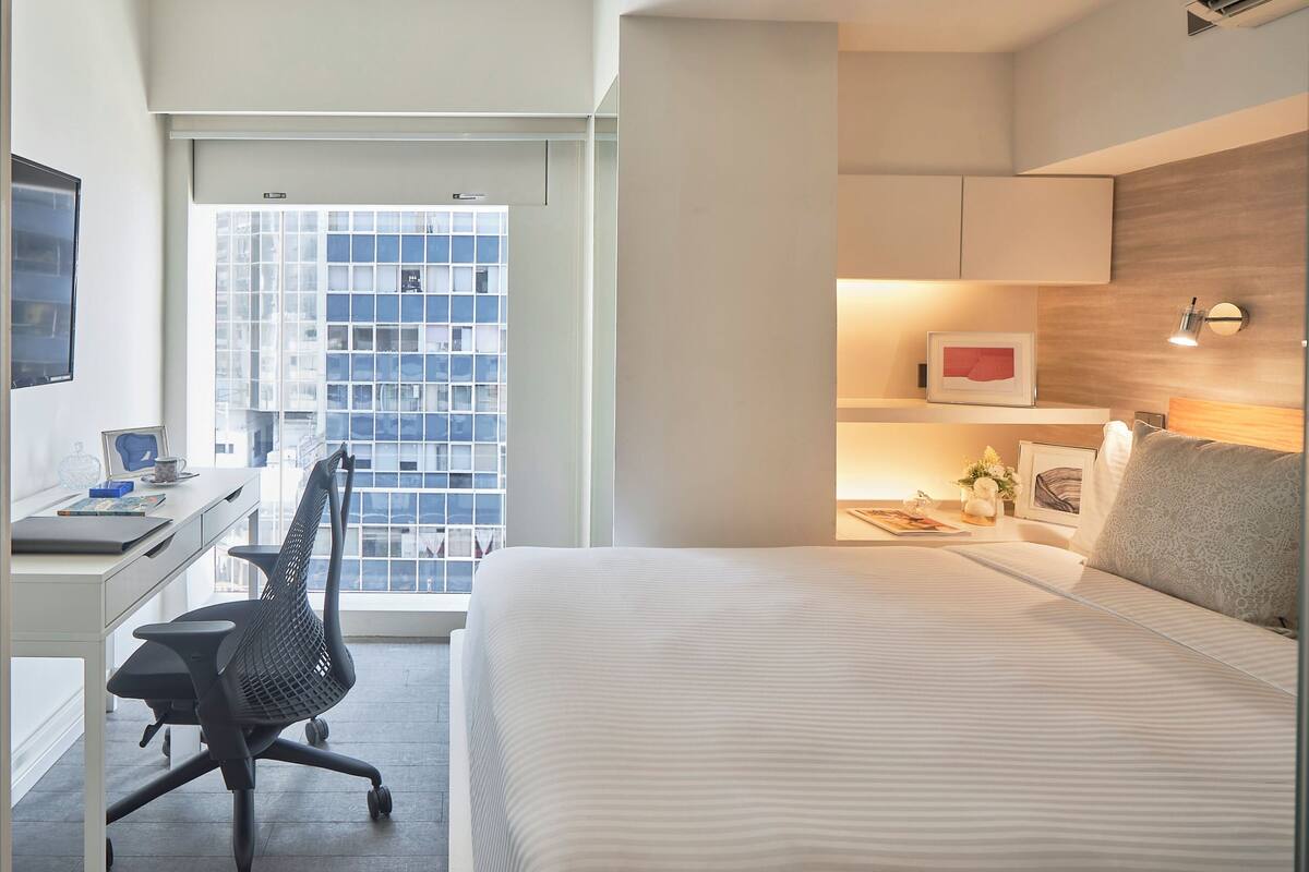 A modern hotel room with a large bed, a desk with a chair, a window with a city view, and a bedside table with a lamp.