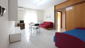 A modern, well-lit room with a flat-screen TV, a dining table with chairs, a red sofa, a blue bed, and a wooden wardrobe. - 1 bedroom beautiful apartment in Parco dei Tigli (Parco dei Tigli)