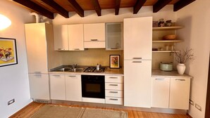 Une cuisine compacte avec des meubles blancs, une cuisinière à gaz, un évier et un petit four. Le plafond est orné de poutres en bois et le mur derrière l’évier est recouvert de carreaux.