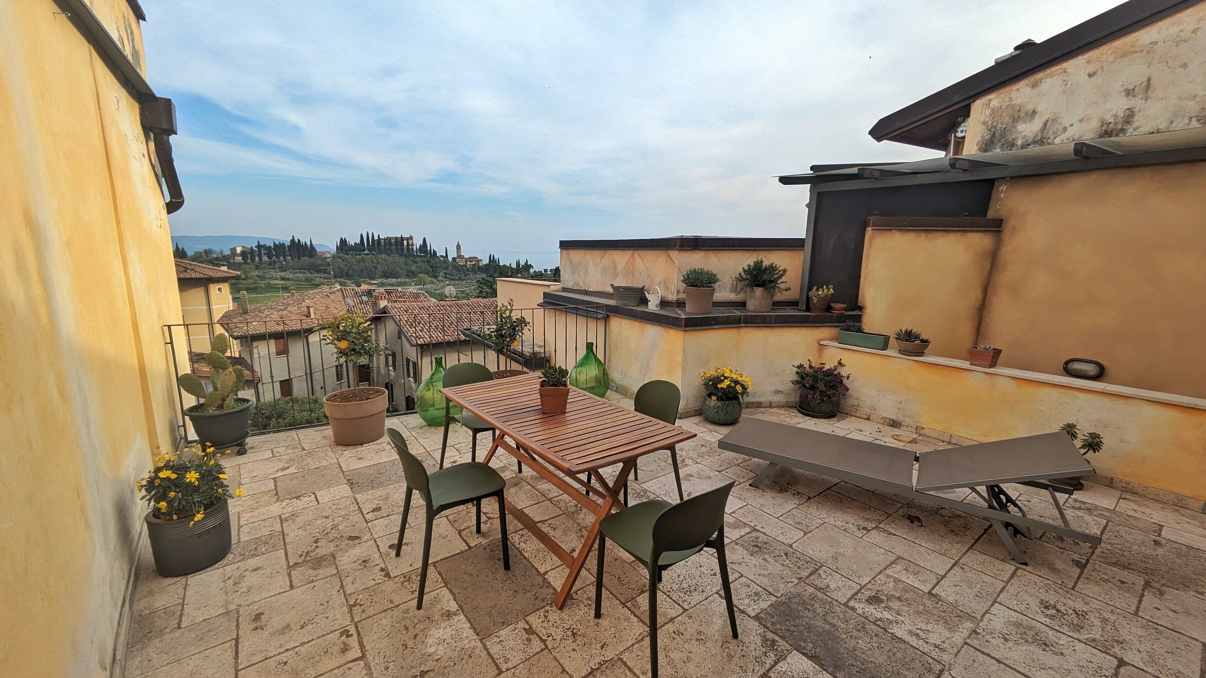 A rooftop terrace with a wooden table and chairs, surrounded by potted plants and a view of the countryside.