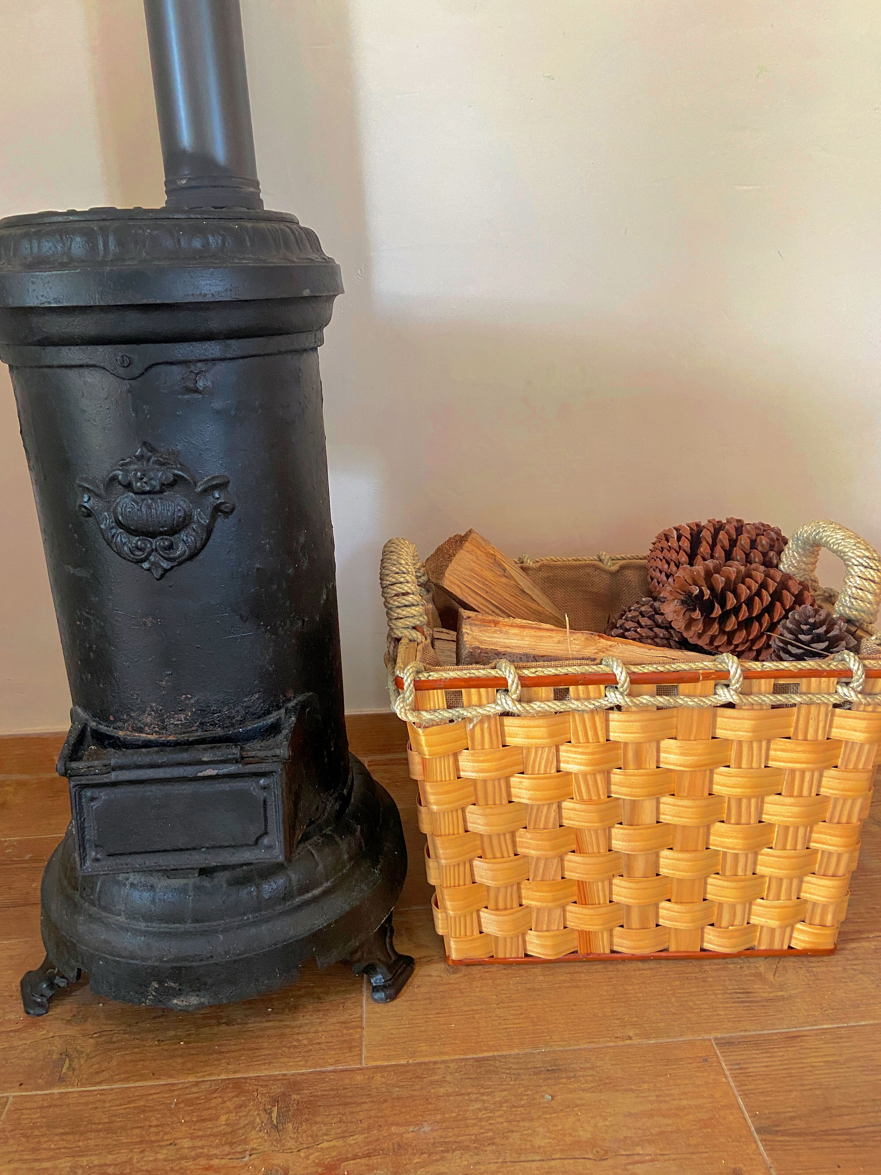 A black wood-burning stove with a decorative emblem, a wicker basket with pinecones, and a wooden floor.
