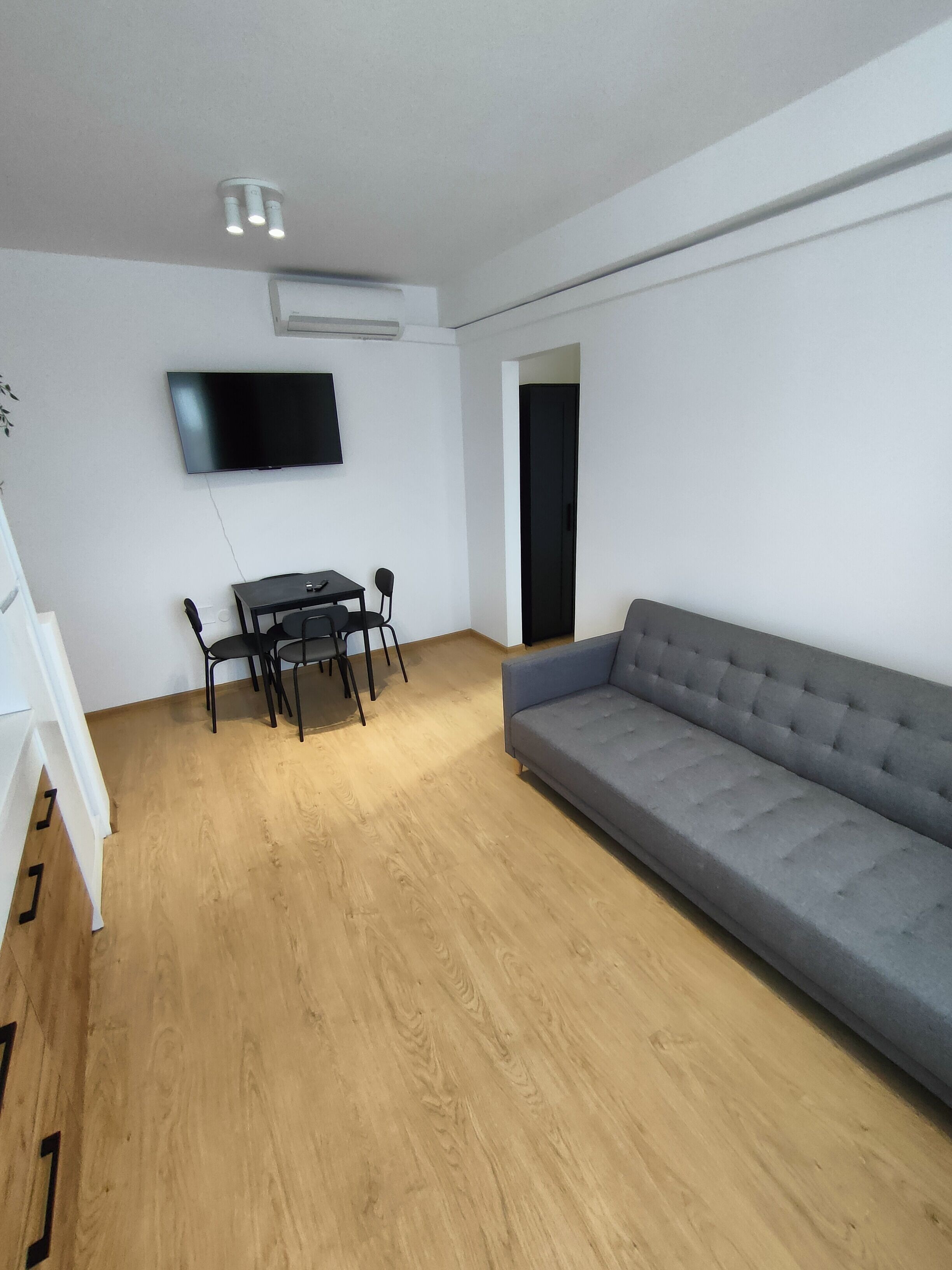 A modern living room with a grey sofa, a dining table with chairs, and a flat-screen TV mounted on the wall.