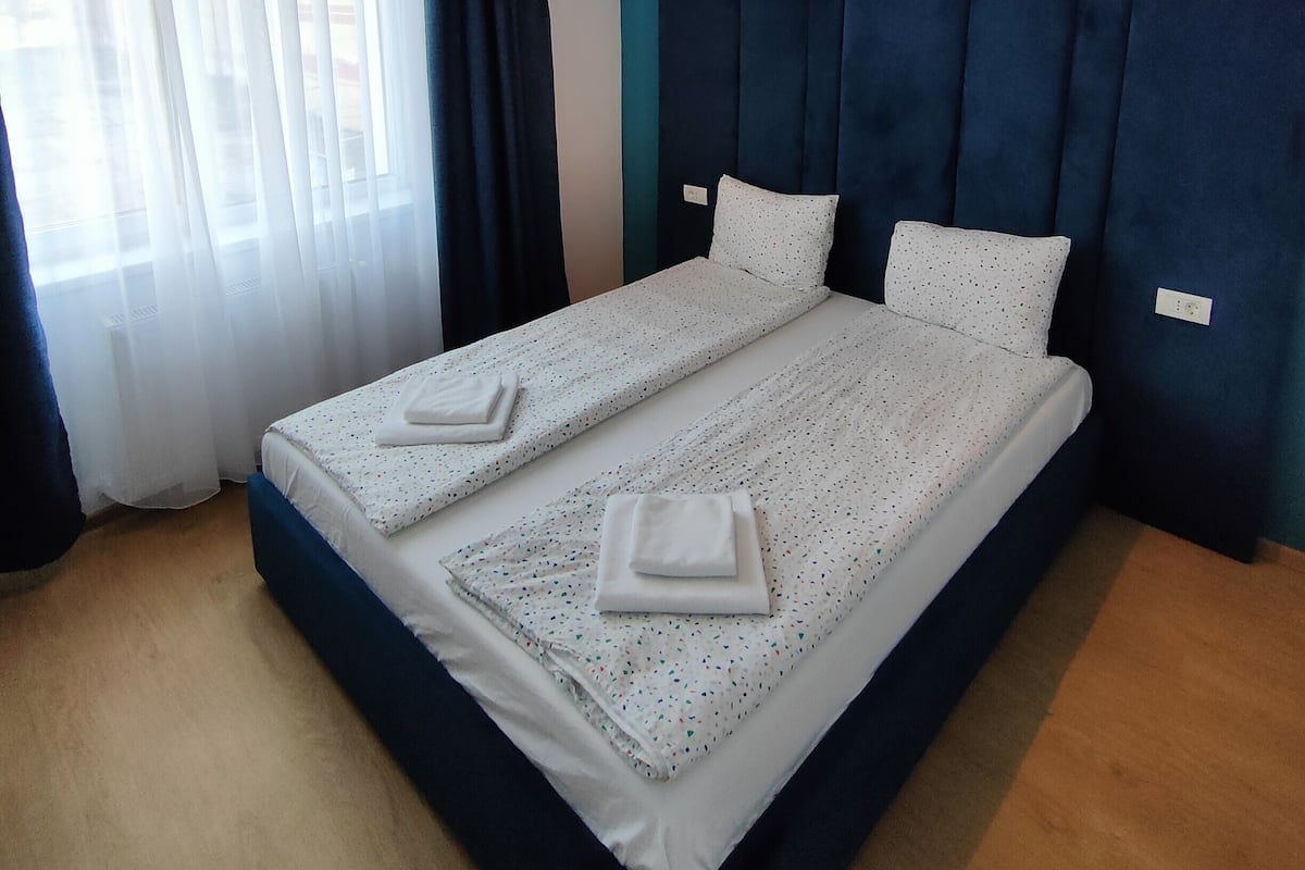 A double bed with white linens and a blue headboard, a window with sheer curtains, and a ceiling light fixture.