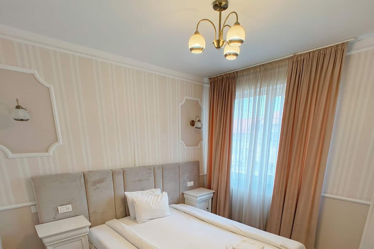 A neatly arranged hotel room with a large bed, bedside tables, a nightstand, a ceiling light fixture, and a window with curtains.