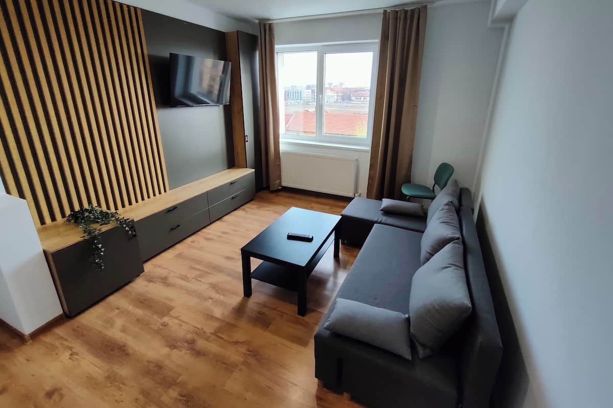 A modern living room with a sofa, a coffee table, a TV mounted on a wooden panel wall, and a window with curtains.