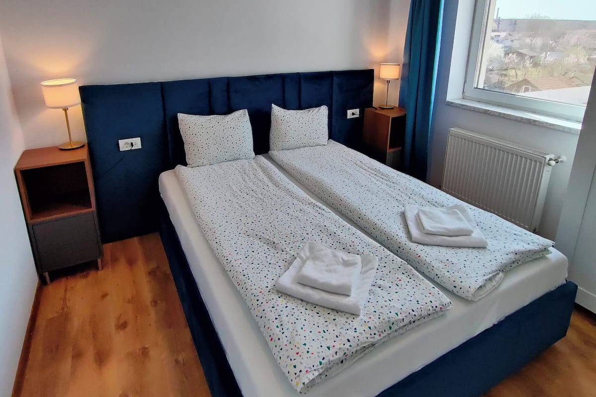 A double bed with a blue headboard, two pillows, and a folded towel on the bed. There is a nightstand with a lamp, a window with a view of buildings, and a radiator.