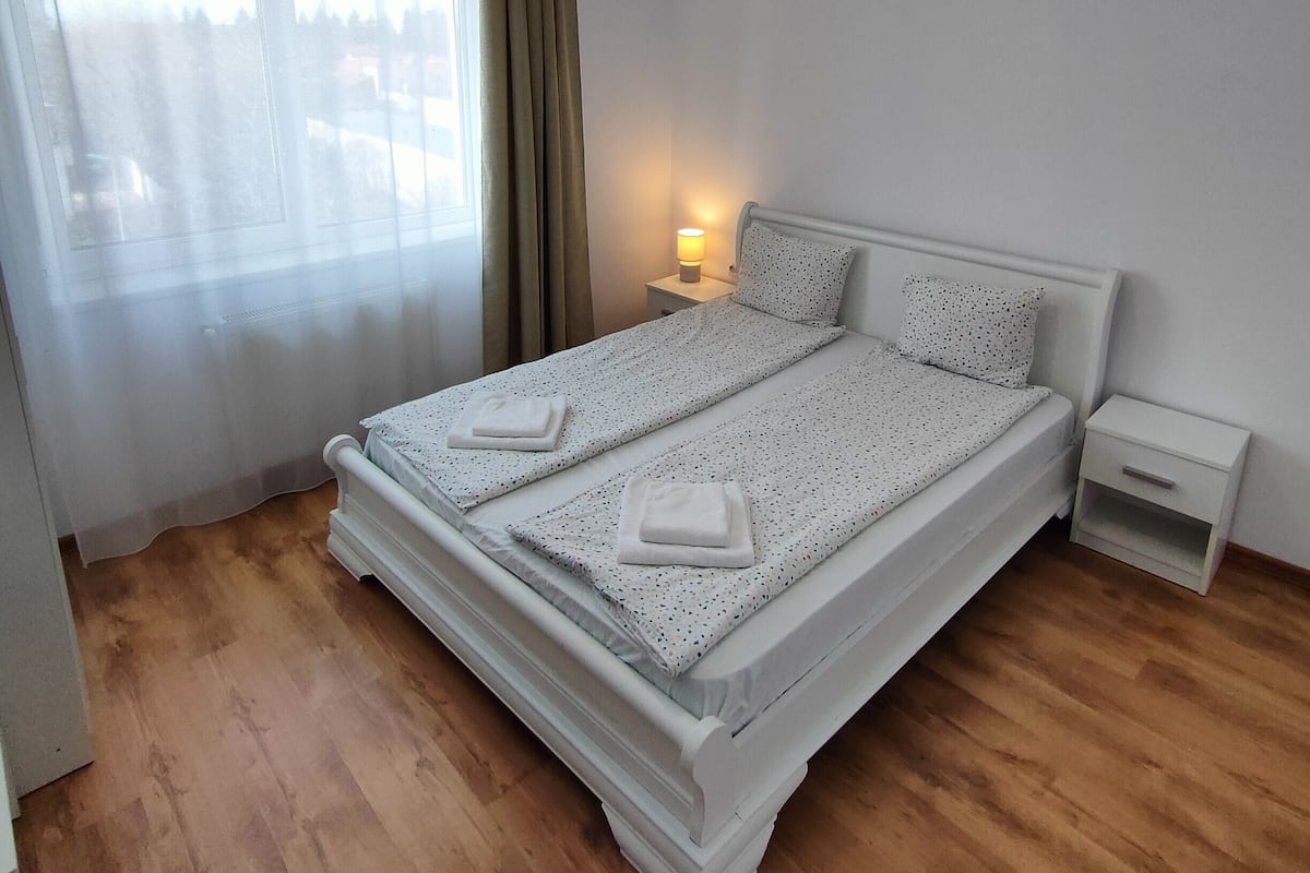 A double bed with a white frame and a patterned bedspread, a bedside table with a lamp, a window with sheer curtains, and a wooden floor.