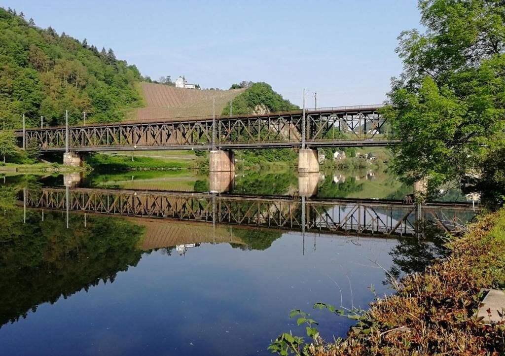 Eine Brücke über ruhigem Wasser, im Hintergrund eine hügelige Landschaft.