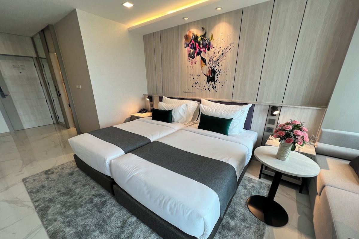 A modern hotel room with a large bed, two bedside tables, a small round table with a vase of flowers, and a decorative wall piece.