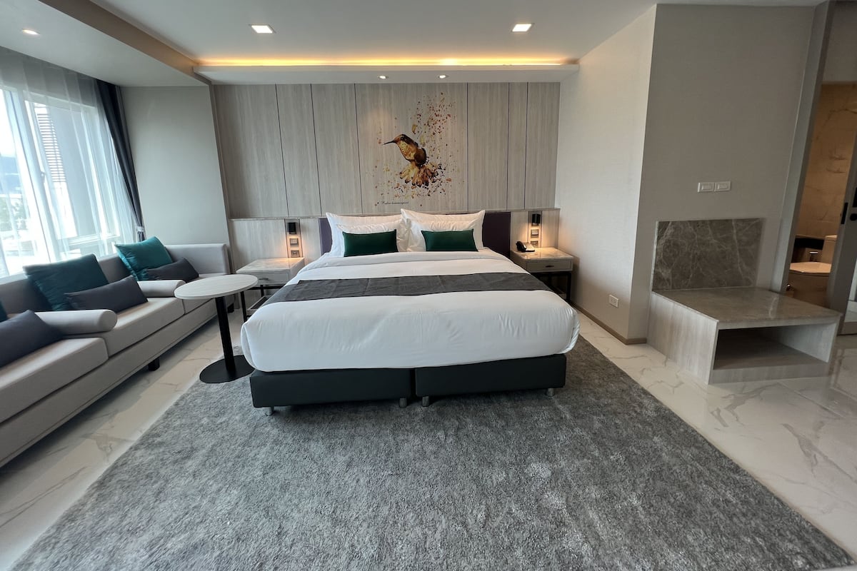 A modern hotel room with a large bed, a sofa, a small table, and a nightstand with a lamp.