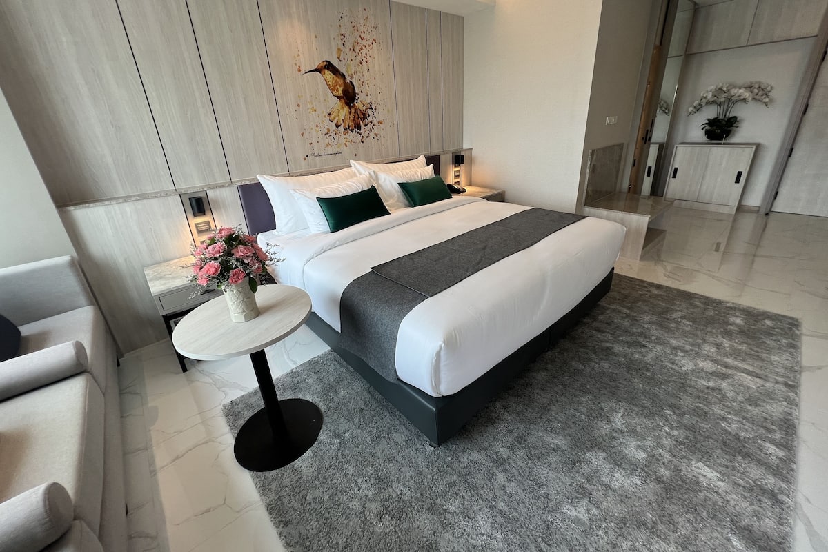 A modern hotel bedroom with a large bed, a bedside table with a vase of flowers, a round side table, and a large mirror.