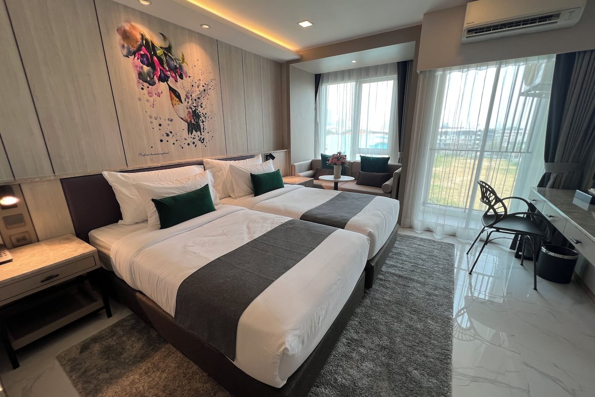 A modern hotel room with two beds, a sofa, a desk, and a large window offering a view of the outdoors.