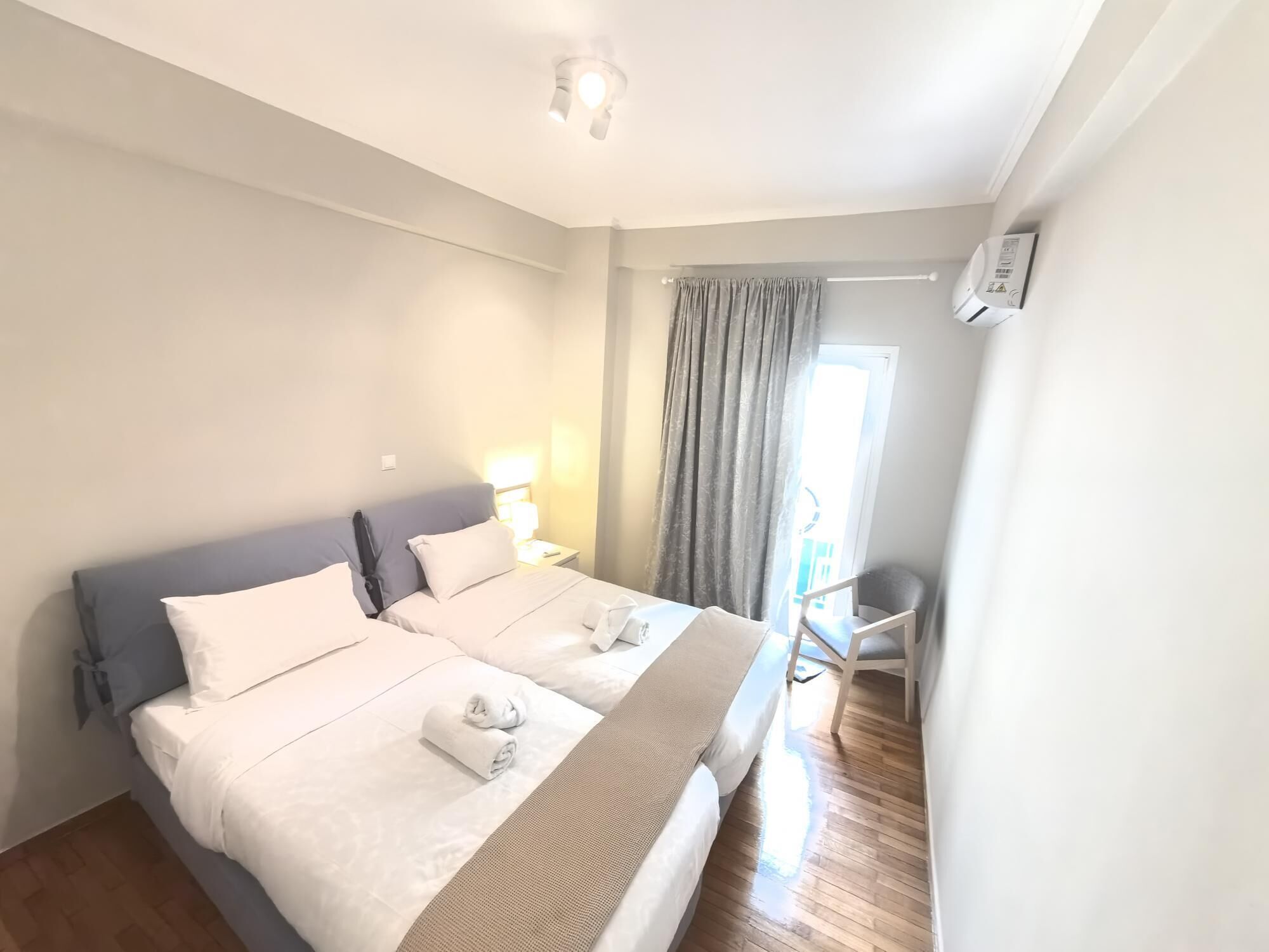 A double bed with white linens, a bedside table with a lamp, a chair, and a window with curtains.