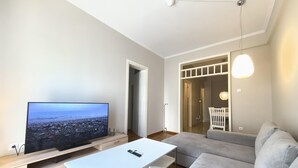 A modern living room with a flat-screen TV, a white coffee table, a grey sofa, and a dining area with a white chair. - Dionysos - Acropolis Residence Apartment (Athens)
