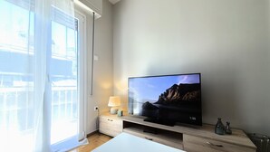 A modern living room with a flat-screen TV on a wooden stand, a white table, and a window with sheer curtains. - Dionysos - Acropolis Residence Apartment (Athens)