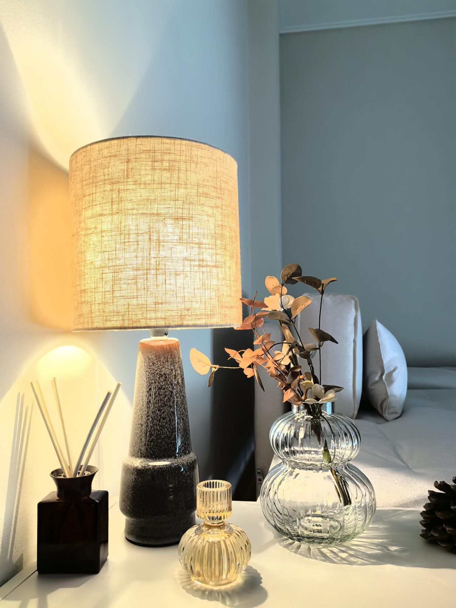 A bedside table with a lamp, a vase with dried flowers, and a candle holder.