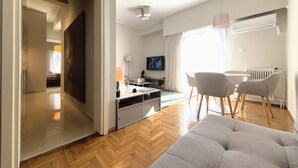 Un salon moderne avec un parquet en chevrons, un canapé gris, un coin repas avec une table et des chaises, un téléviseur fixé au mur et une grande fenêtre laissant entrer la lumière naturelle.