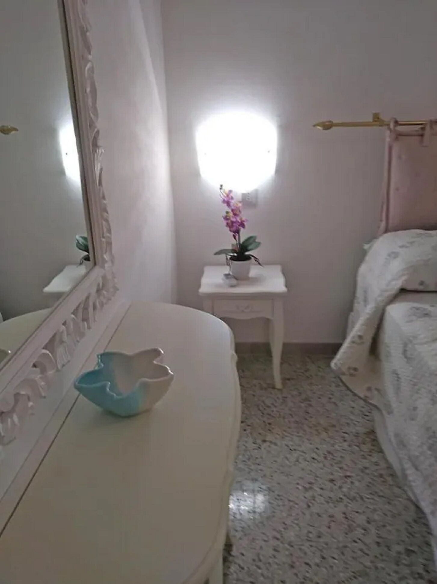 A bedroom with a bed, a bedside table with a flower vase, a mirror, and a light fixture on the wall.