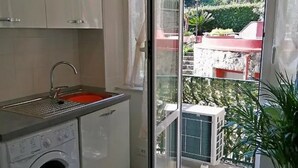 A compact kitchen with a washing machine, a sink, and a glass door leading to a balcony with a view of greenery and buildings.