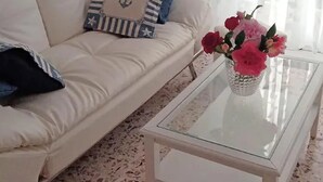 A white sofa with a striped pillow, a glass coffee table with a vase of flowers, and a window with sheer curtains.