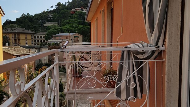 A balcony with ornate white railings, a tiled floor, and a view of a hillside with buildings.