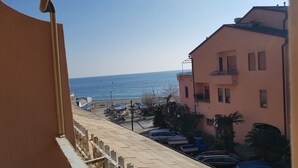 A balcony with a view of the sea, parked cars, and a building with balconies.