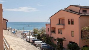 A coastal street with parked cars, a building with balconies, and a view of the sea.