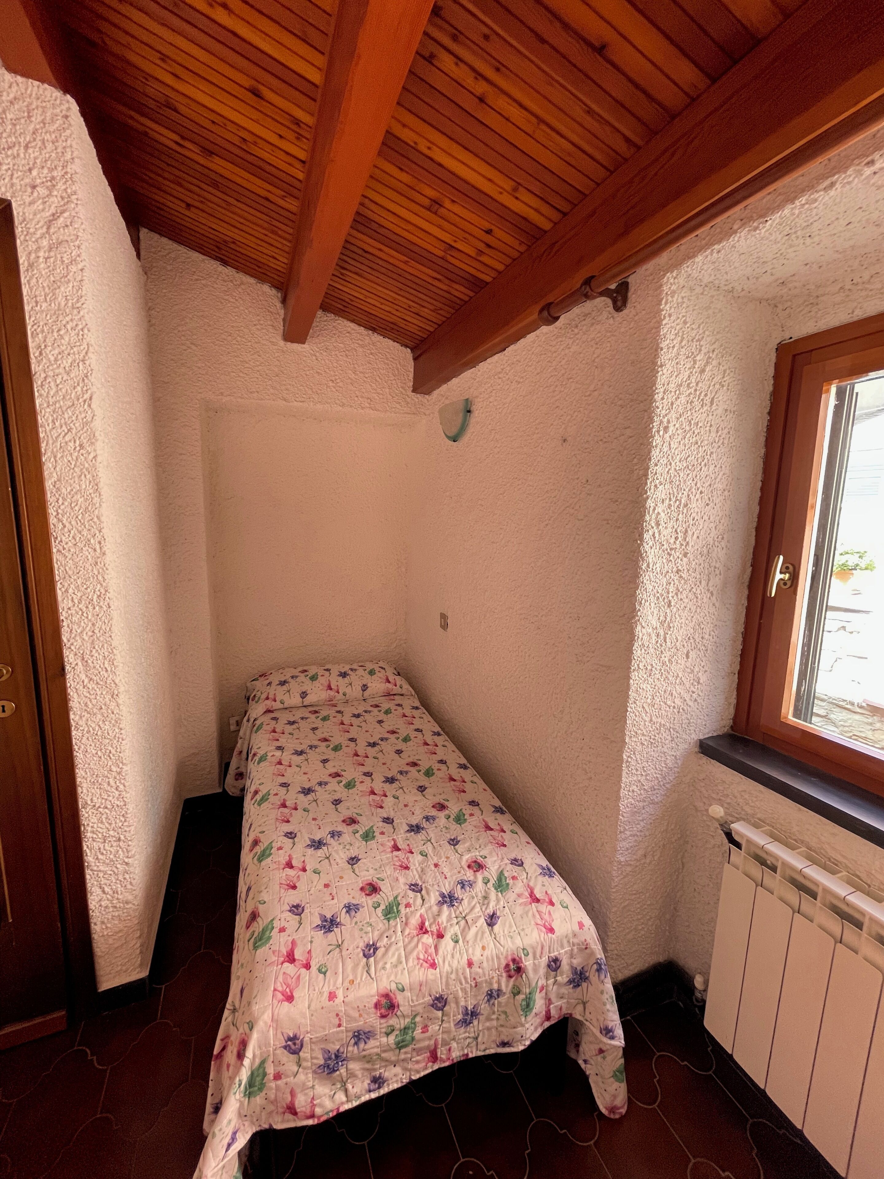 Ein kleiner, gemütlicher Raum mit Holzbalkendecke, einem Einzelbett mit floralem Überwurf, einem Fenster mit Blick und einer Heizung.