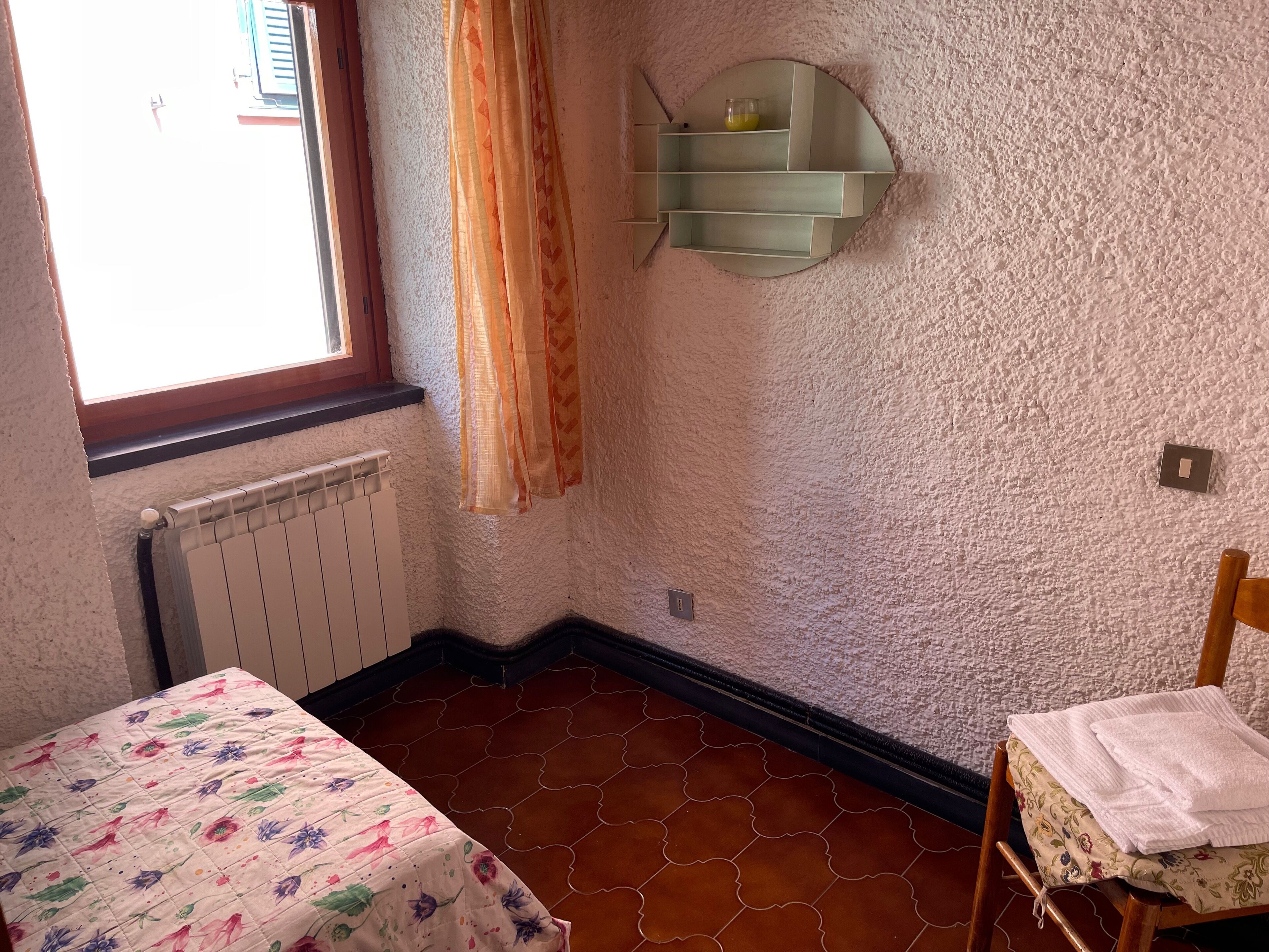 Une pièce avec un radiateur, un lit recouvert d’une couvre-lit à motifs floraux, une chaise avec une serviette, une fenêtre avec des rideaux et une étagère fixée au mur.