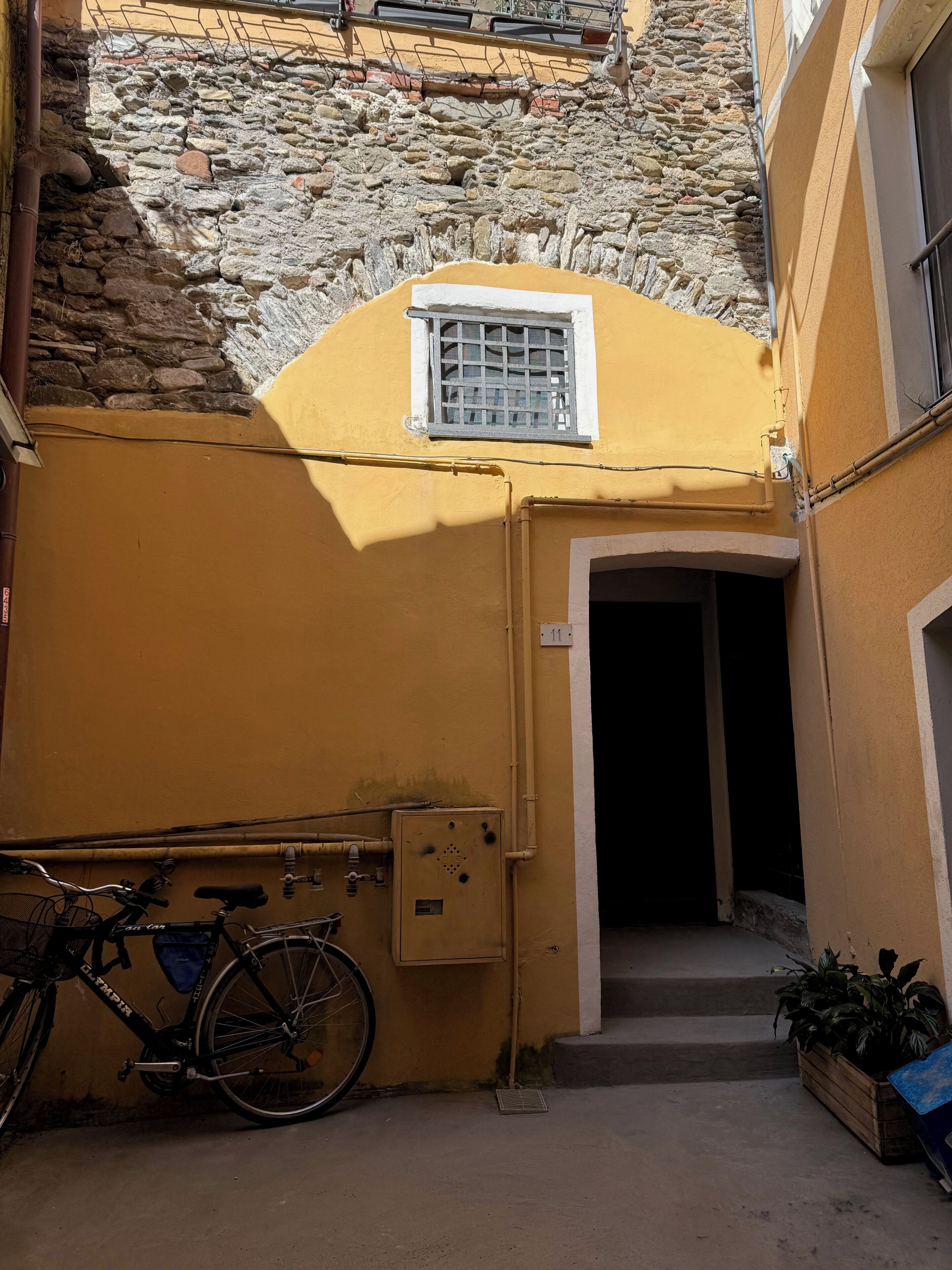 A narrow alleyway with a yellow wall, a stone wall, and a metal gate. A bicycle is parked against the wall, and there is a small window with a metal grille.
