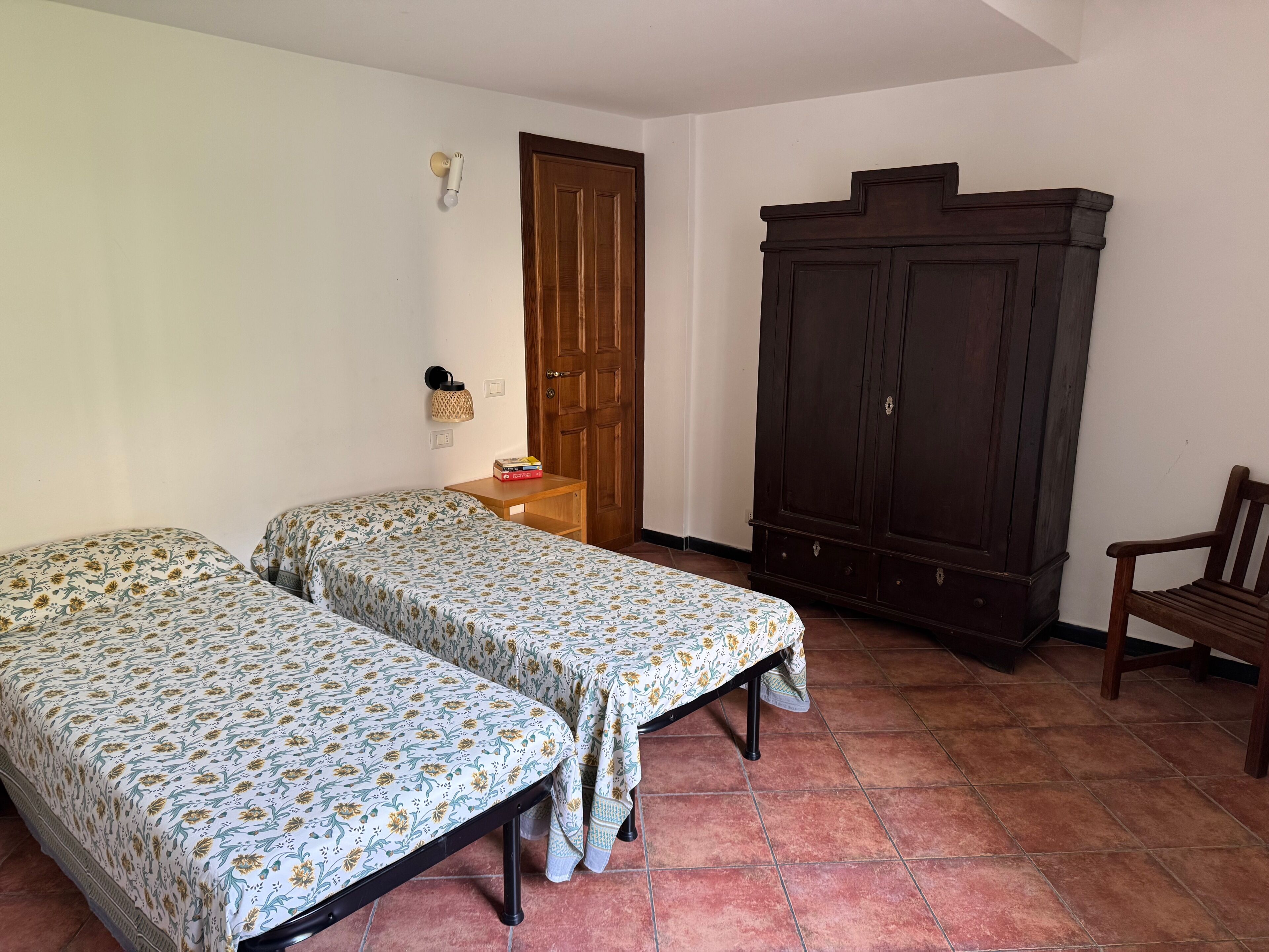 Une chambre avec deux lits simples, une armoire en bois, une petite table en bois avec une lampe et un banc.